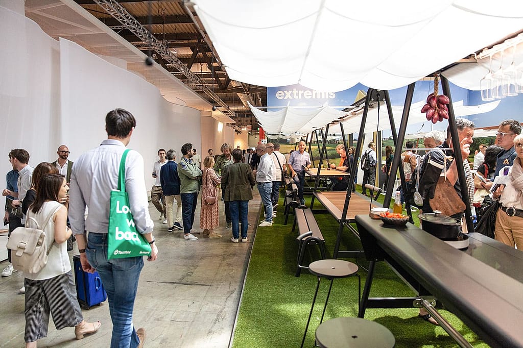 Vue complète du stand montrant l’installation modulaire AMAi disposée comme une plate-forme de jardin avec de longues tables, des tabourets tiki et un auvent en tissu blanc au-dessus.