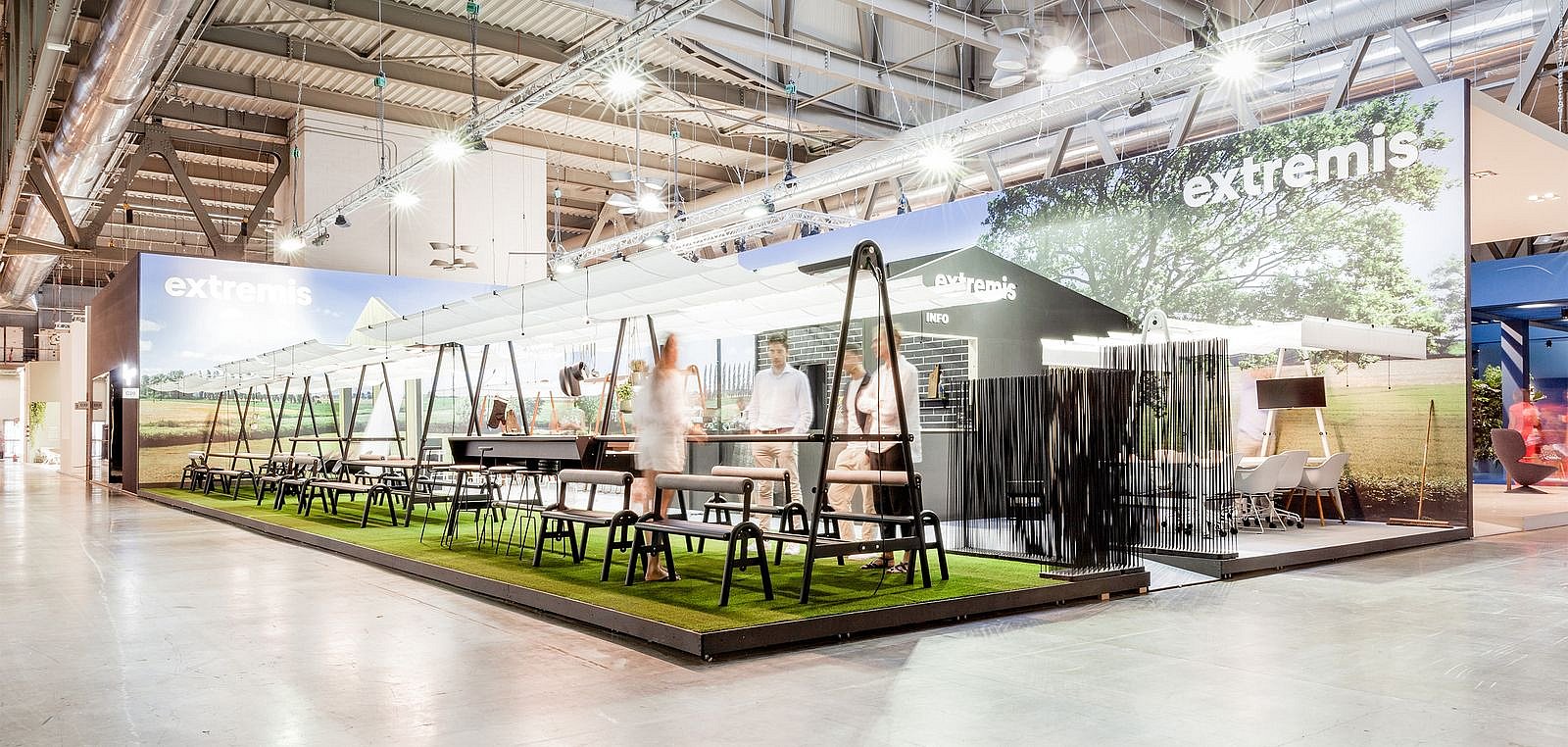 Vue dégagée du stand de la foire AMAi avec de longues tables à cadre noir, des tabourets, du gazon artificiel et de grands panneaux d’ombrage blancs créant une atmosphère de fête dans le jardin.
