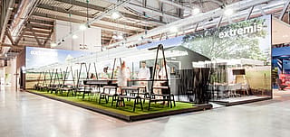 Vue dégagée du stand de la foire AMAi avec de longues tables à cadre noir, des tabourets, du gazon artificiel et de grands panneaux d’ombrage blancs créant une atmosphère de fête dans le jardin.