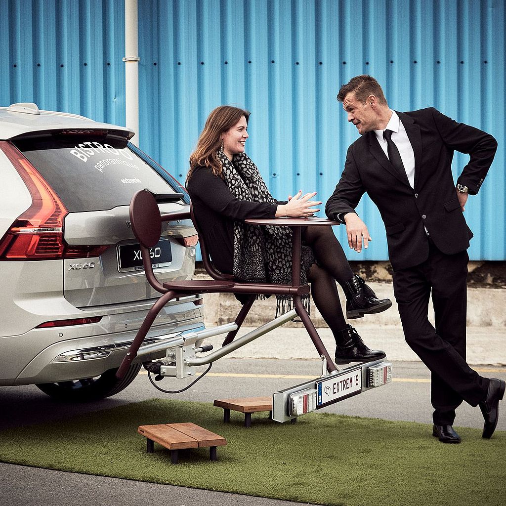Vrouw zittend op een compacte Bistroo zit- en tafelunit, gemonteerd achterin een auto, in gesprek met een man in een zwart pak voor een felblauwe golfplaten muur.