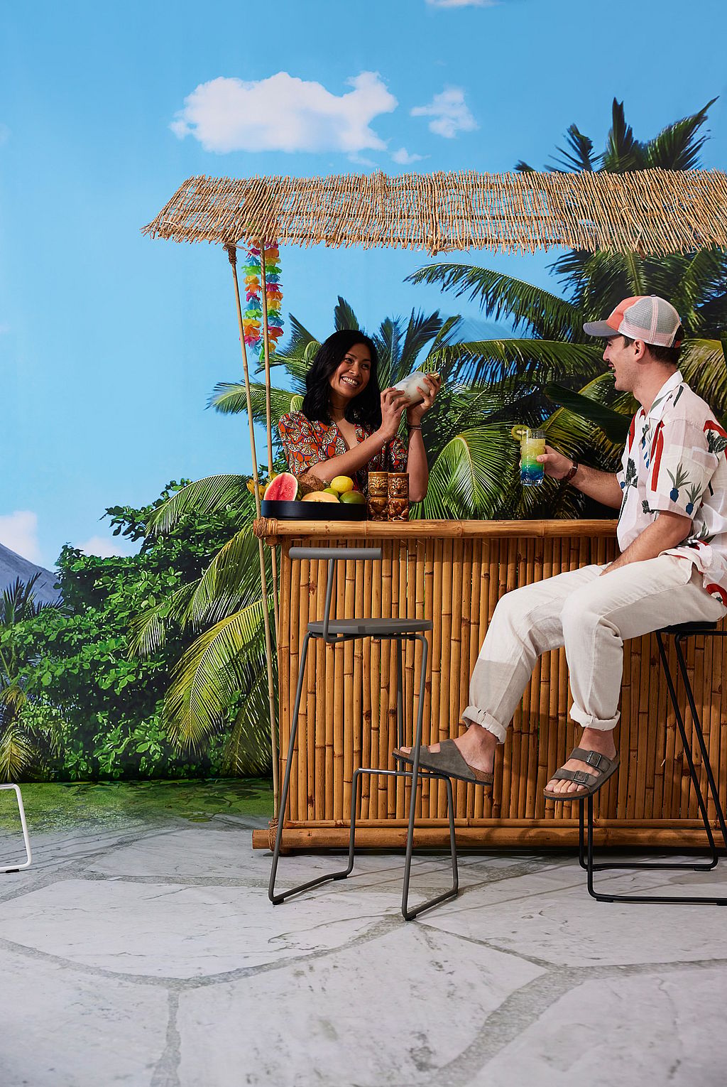Eine Frau hinter einer Bambus-Tiki-Bar trinkt einen Cocktail, während ein Mann auf einem schwarzen Tiki-Hocker sitzt und ein Getränk hält - mit Palmblättern und einer Strohdecke, die eine festliche Bar-Atmosphäre schafft.