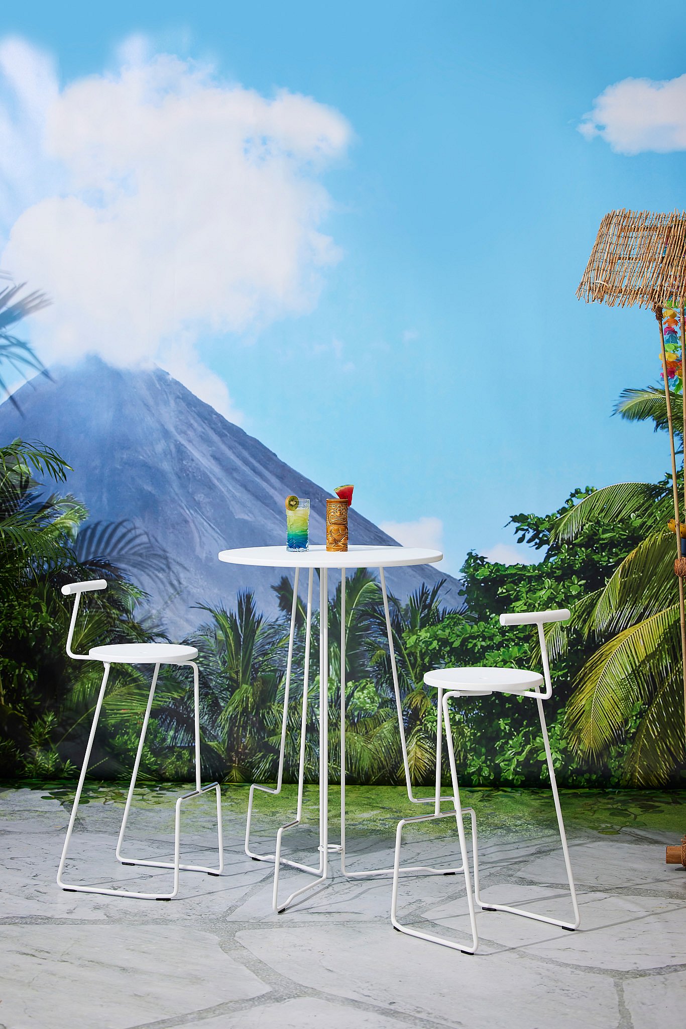 White Tiki, stehender Tisch mit zwei aufeinander abgestimmten Hockern vor einer tropischen Wandmalerei, mit bunten Cocktails auf dem Tisch und einem im Hintergrund aufsteigenden Vulkan.