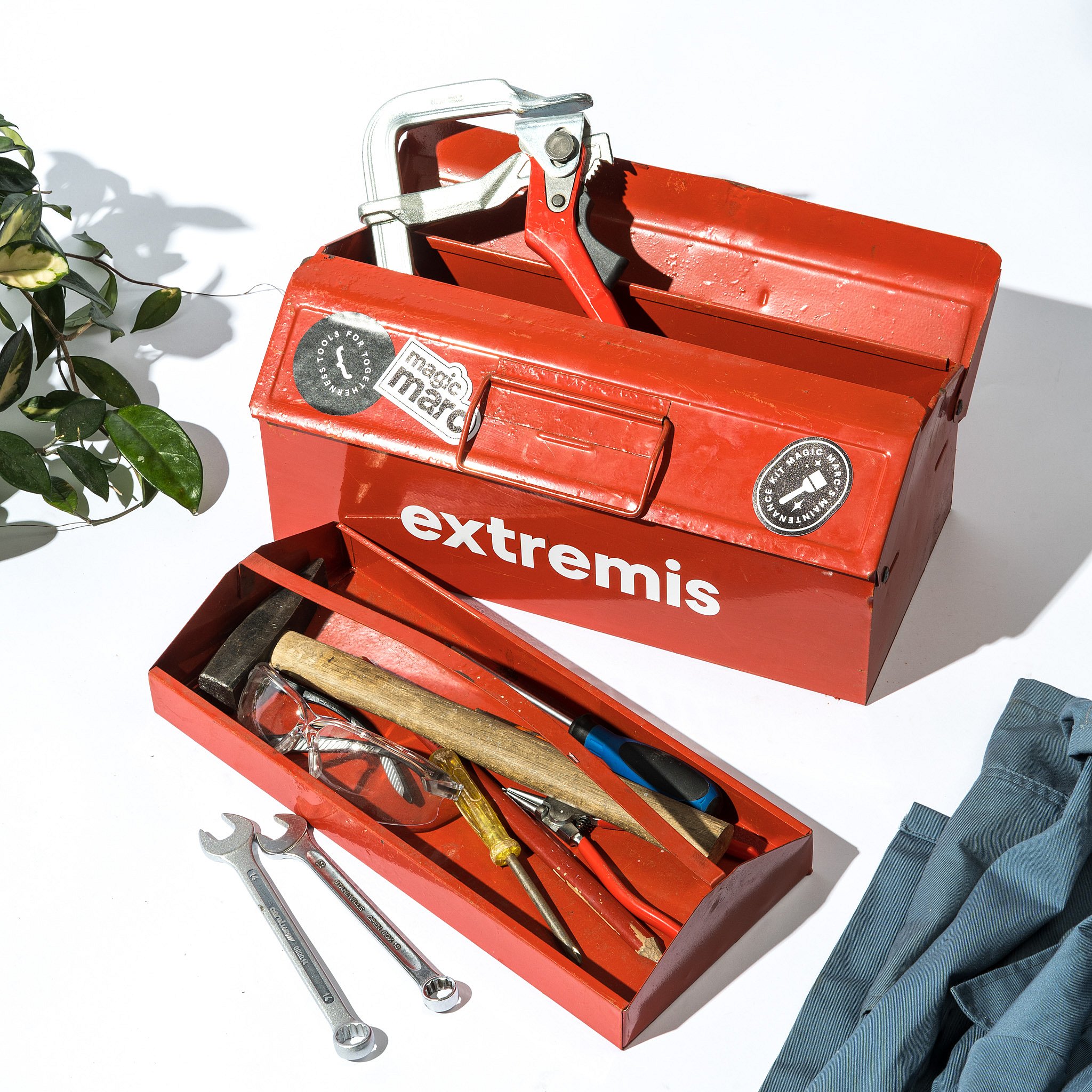 Open red metal toolbox with Extremis branding, filled with hand tools including a clamp, hammer, screwdrivers, pliers, safety glasses, and wrenches on a white surface.