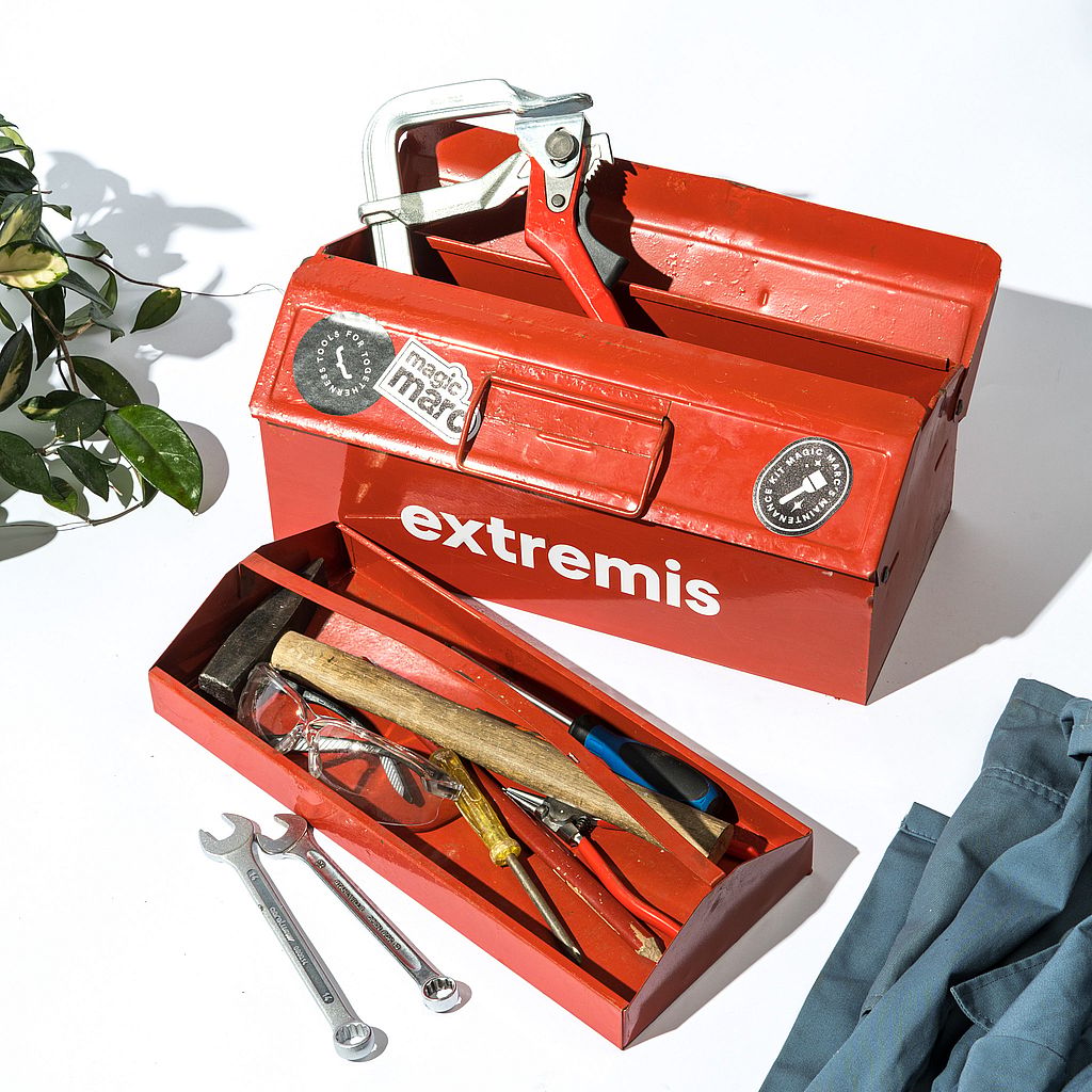 Open red metal toolbox with Extremis branding, filled with hand tools including a clamp, hammer, screwdrivers, pliers, safety glasses, and wrenches on a white surface.