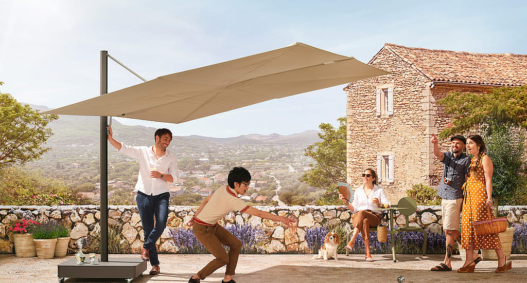 Mensen spelen jeu de boules op een zonnig terras onder een grote beige parasol, met een stenen huis en panoramisch uitzicht op de heuvel op de achtergrond.