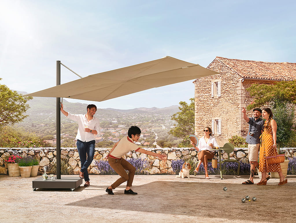 Menschen spielen Pétanque auf einer sonnigen Terrasse unter einem großen beigen Sonnenschirm, mit einem Steinhaus und Panoramablick auf den Hang im Hintergrund.