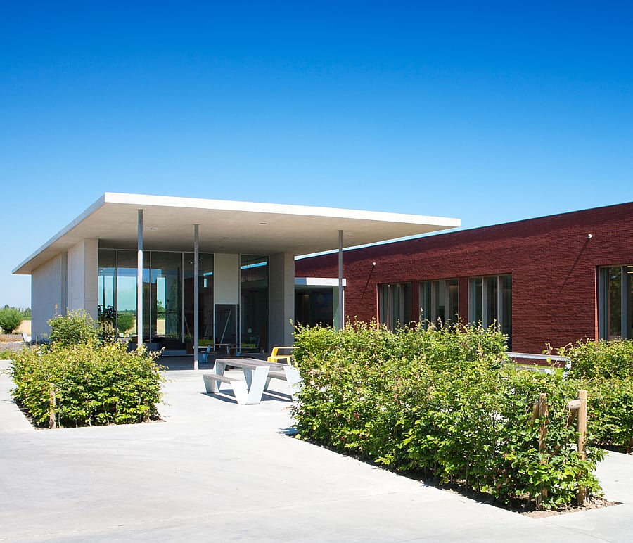 Modern Extremis-gebouw met een witte luifelingang, rode bakstenen gevel, aangelegde heggen en tuinmeubilair onder een helderblauwe hemel.