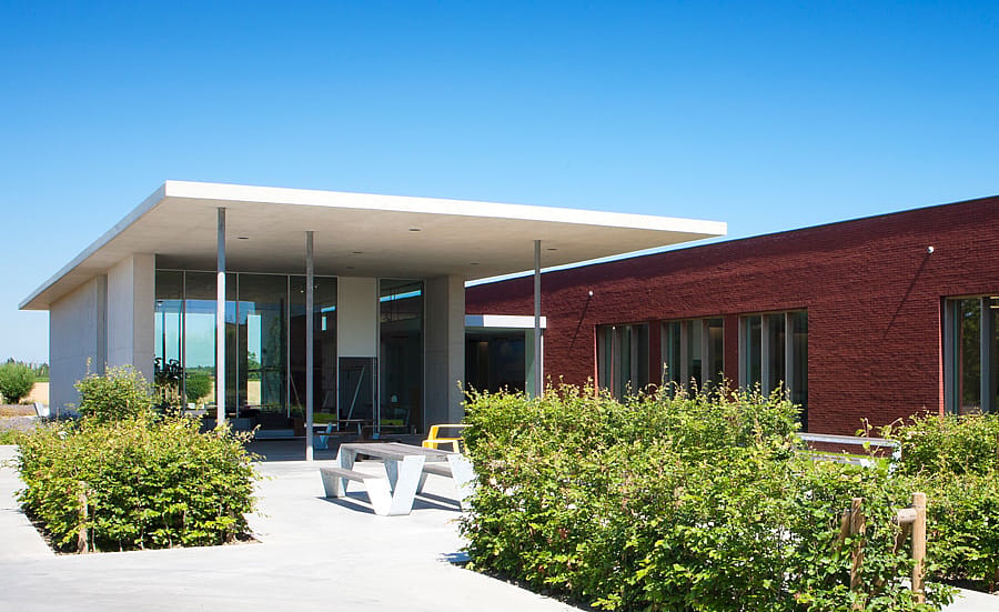 Modern Extremis-gebouw met een witte luifelingang, rode bakstenen gevel, aangelegde heggen en tuinmeubilair onder een helderblauwe hemel.