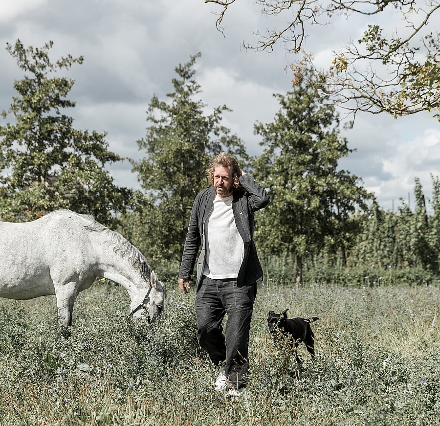 Dirk Wynants loopt door een weiland met een wit paard dat naast hem graast en een zwarte hond die in de buurt volgt onder een bewolkte hemel.