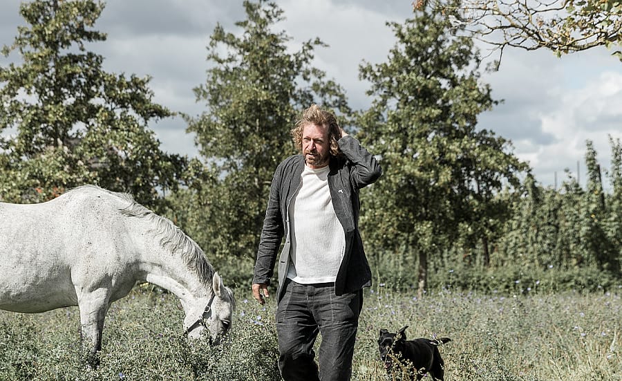 Dirk Wynants loopt door een weiland met een wit paard dat naast hem graast en een zwarte hond die in de buurt volgt onder een bewolkte hemel.