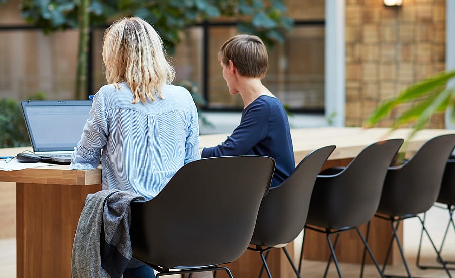 Twee mensen zaten aan een lange houten tafel met zwarte kapiteinsstoelen en werkten samen aan een laptop in een rustige binnenruimte met planten en grote ramen.