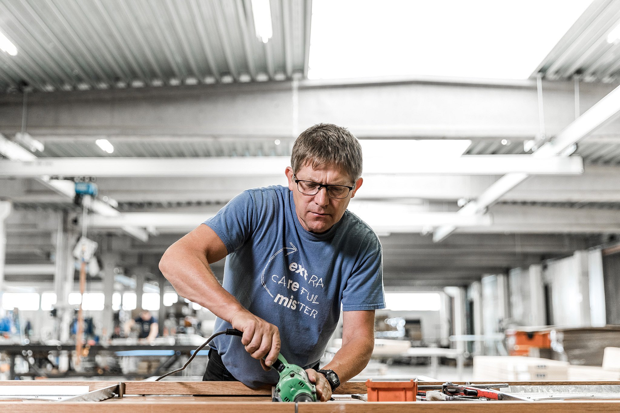 Marc gebruikt een elektrisch gereedschap op een houten meubelcomponent in de werkplaats en toont praktische productie bij Extremis.
