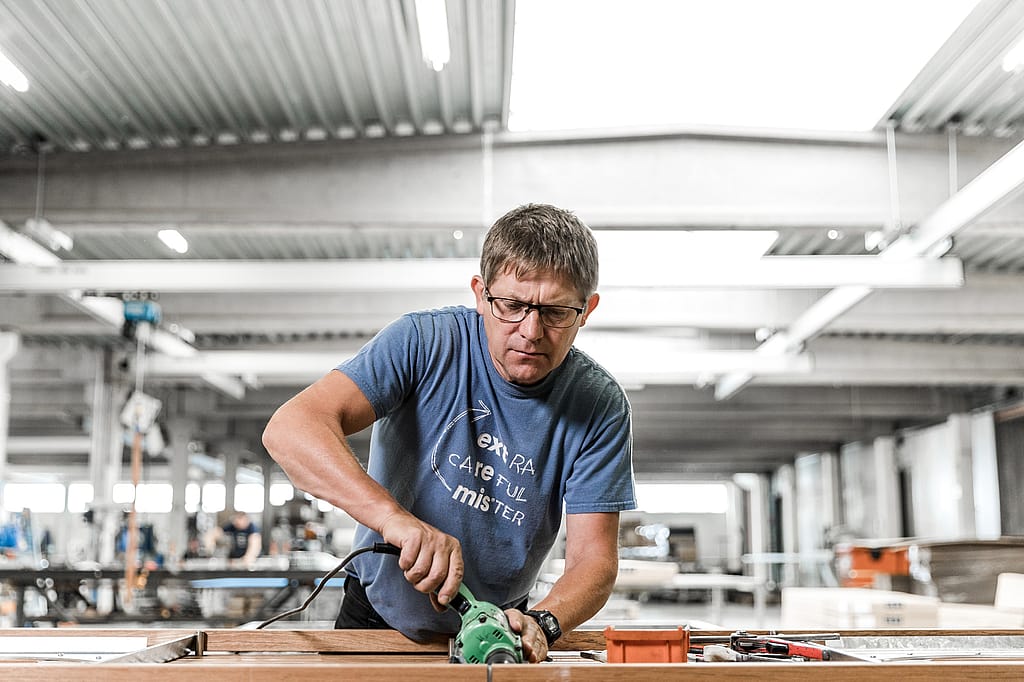 Marc gebruikt een elektrisch gereedschap op een houten meubelcomponent in de werkplaats en toont praktische productie bij Extremis.