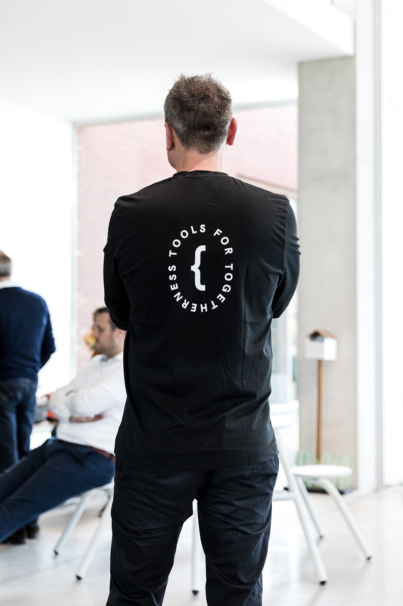 Persoon die binnenshuis staat met een zwart shirt met de tekst ‘Gereedschap voor saamhorigheid ’ op de rug, met collega's op de achtergrond wazig.