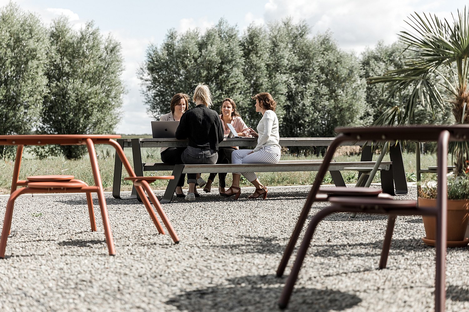 Coworkers zaten samen aan een lange hoppertafel voor op grind en hadden een ontmoeting omringd door bomen en planten.
