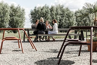 Coworkers zaten samen aan een lange hoppertafel voor op grind en hadden een ontmoeting omringd door bomen en planten.