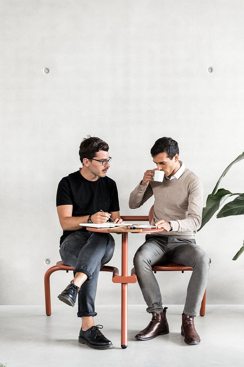 Twee collega's aan een compacte koperen bruine bistrootafel, de ene drinkt koffie en de andere schrijft aantekeningen.