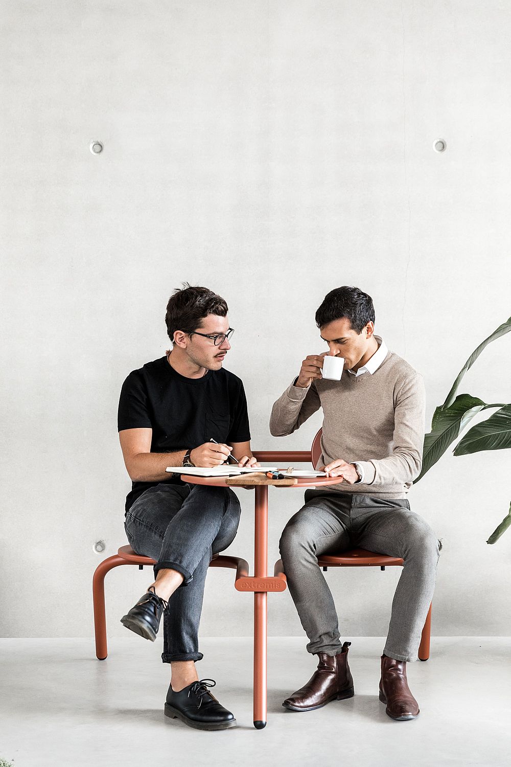 Twee collega's aan een compacte koperen bruine bistrootafel, de ene drinkt koffie en de andere schrijft aantekeningen.