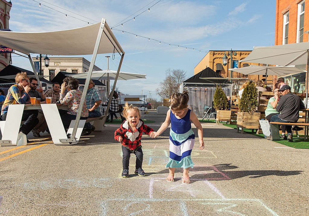 Children playing hopscotch in the middle of a lively pedestrian street, with people dining under shade canopies and parasols in the background.