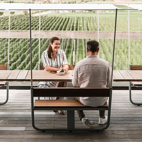 Deux personnes assises l’une en face de l’autre à une table manille avec un auvent ombragé, surplombant des rangées de vignobles depuis une terrasse extérieure.