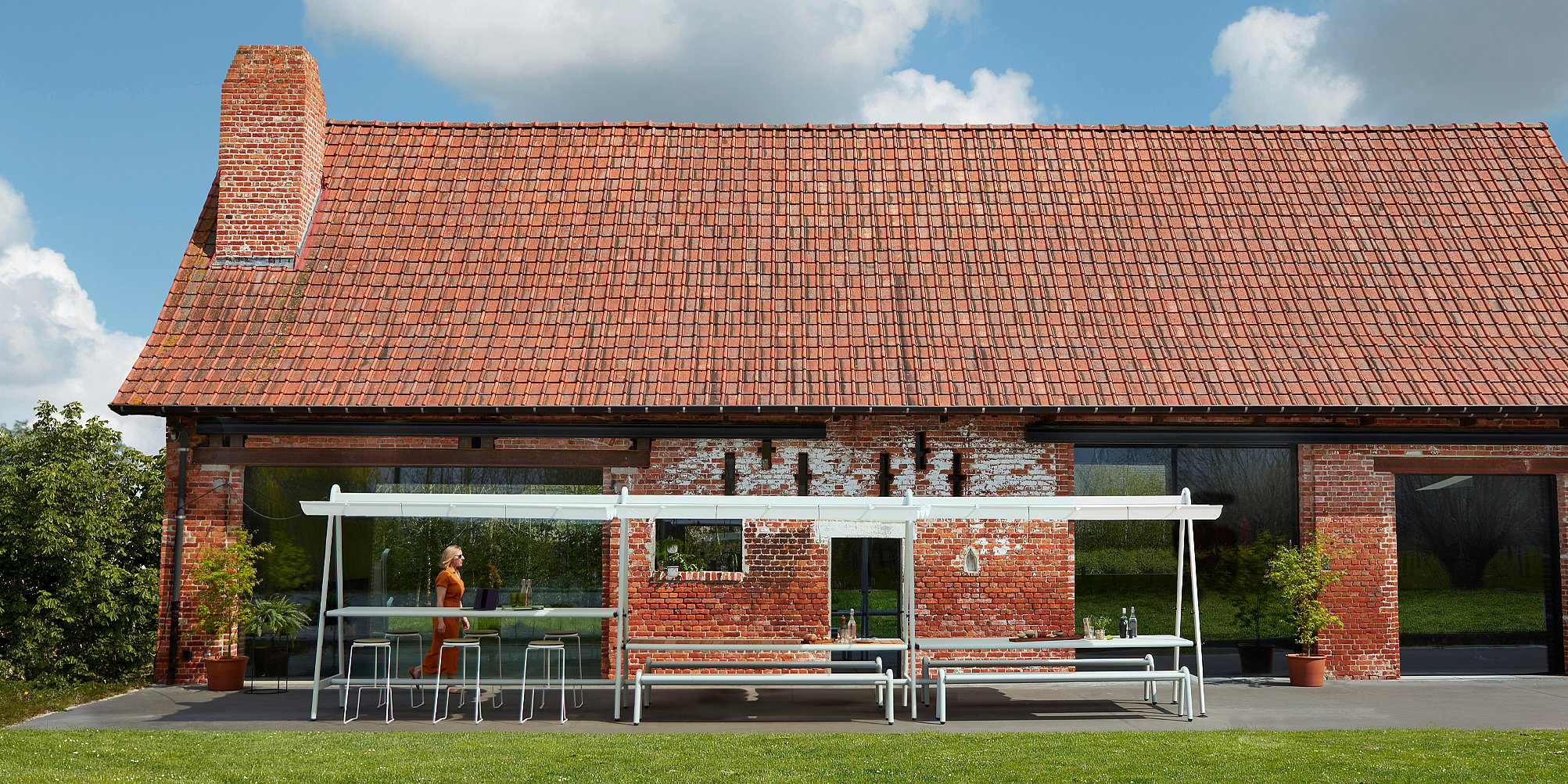 Wide view of multiple AMAi tables connected outside a red brick building, creating a long shared table arrangement on a green lawn.
