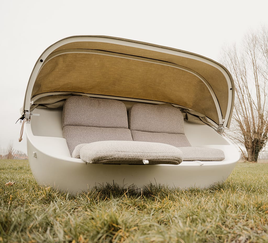 Cremefarbenes Cabrio-Außensofa mit teilweise erhöhtem Softtop, dargestellt als kompakte Zwei-Sitzer-Lounge in einer angelegten Umgebung.