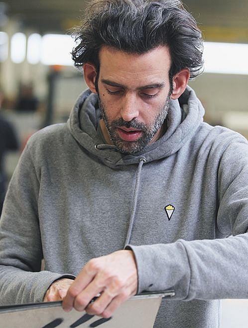 Denis Meyers est assis à une simple table de Picnik rouge sur une palette en bois dans un atelier, peignant à la main des lettres noires sur le plateau.
