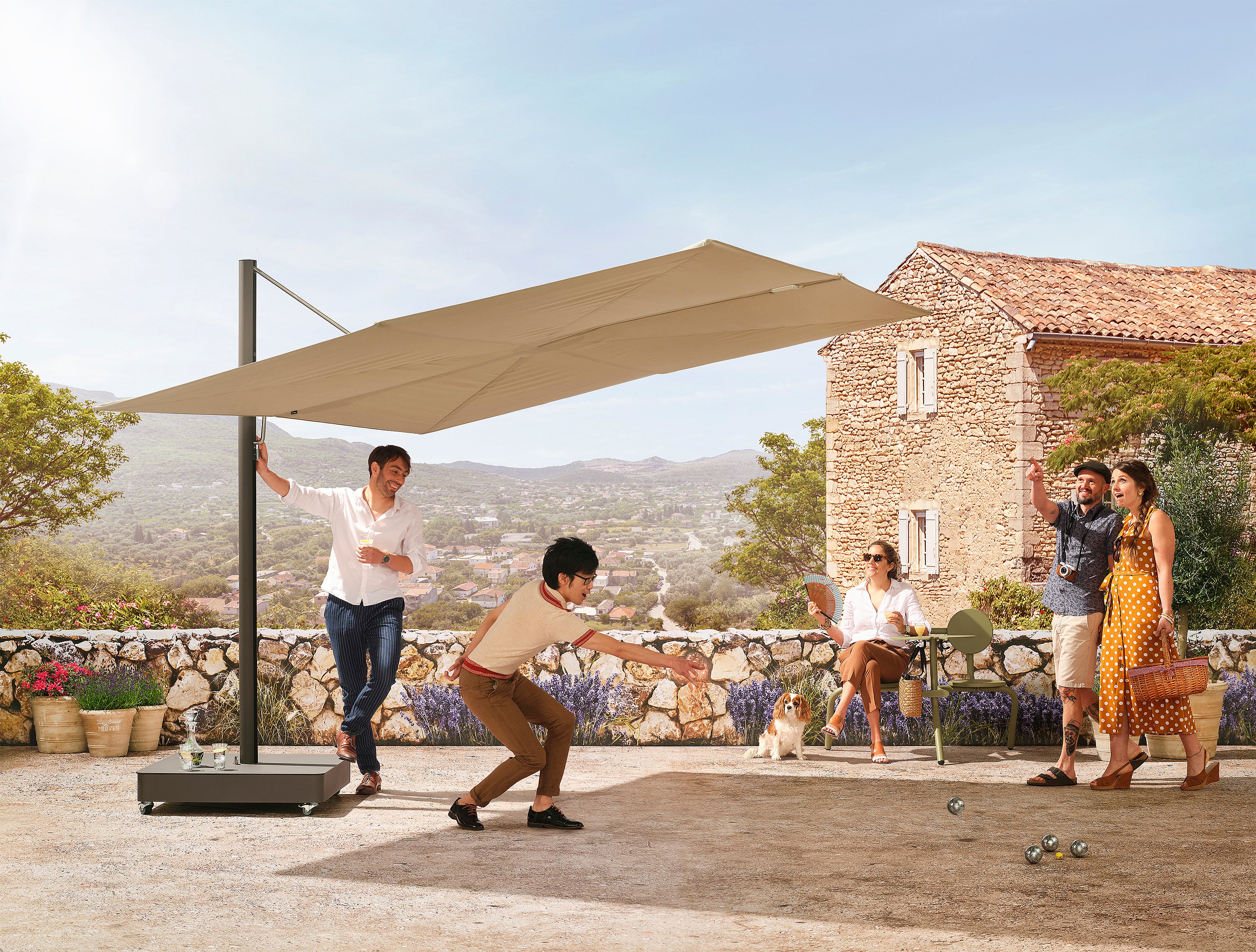 Groupe de personnes jouant à la pétanque sur une terrasse ensoleillée, sous un grand parasol Sensu beige. Derrière eux, une maison en pierre et un panorama sur les collines.