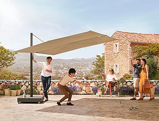 Groupe de personnes jouant à la pétanque sur une terrasse ensoleillée, sous un grand parasol Sensu beige. Derrière eux, une maison en pierre et un panorama sur les collines.
