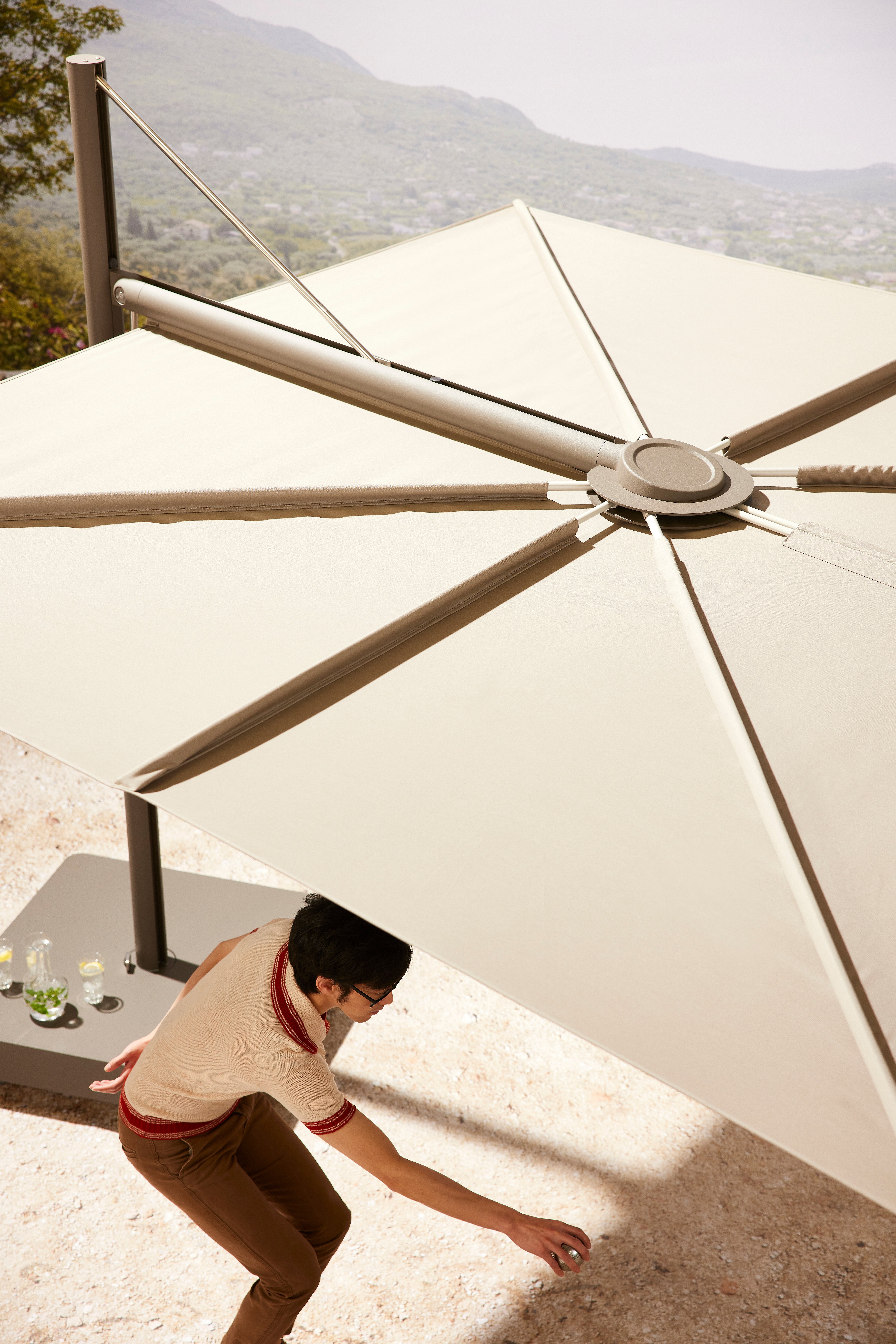 Vue plongeante d'un parasol Sensu beige faisant de l'ombre à une personne jouant à la pétanque. Paysage vallonné en arrière-plan.