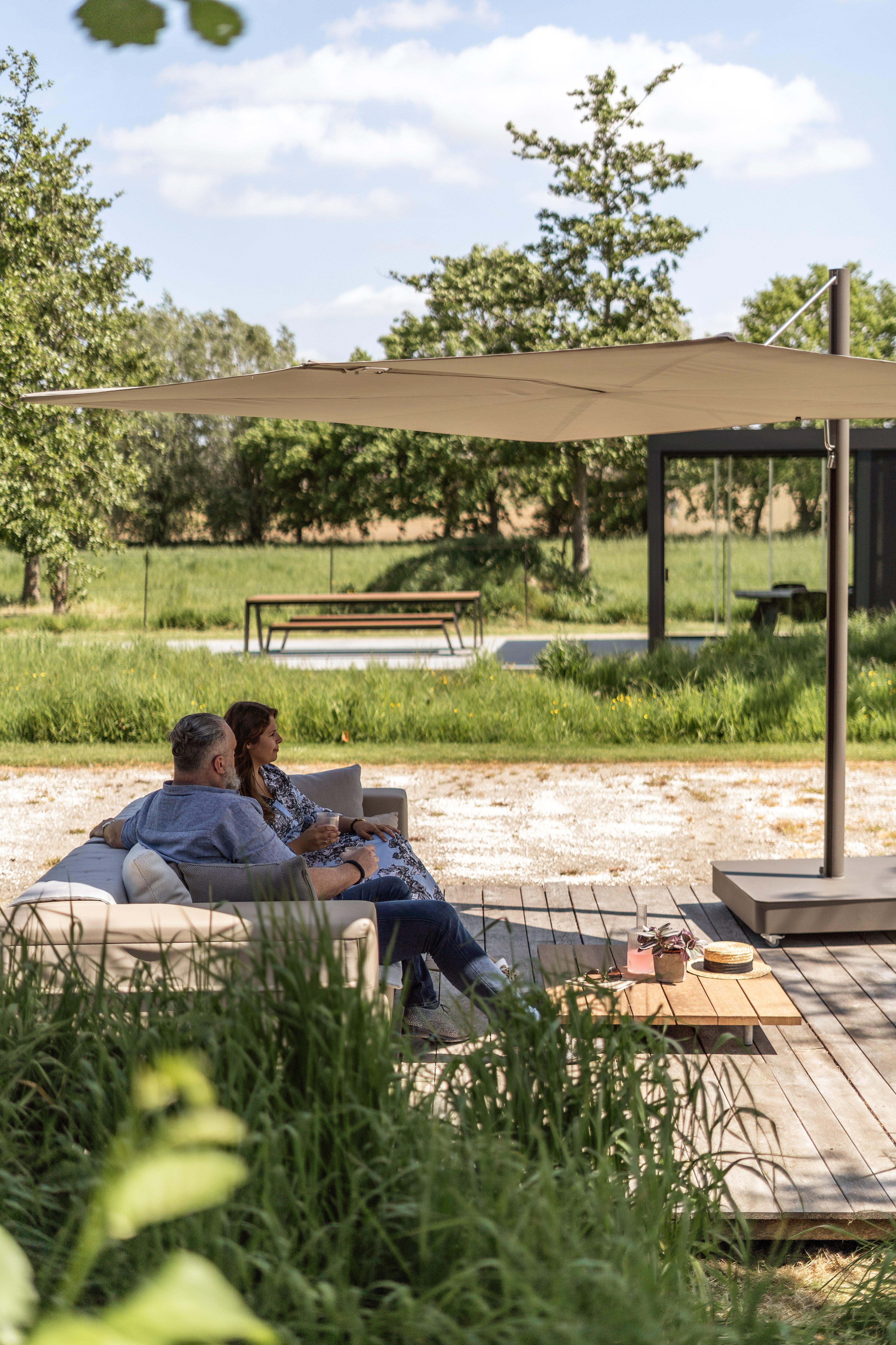 Couple se détendant sur un canapé de jardin sous un grand parasol Sensu beige, dans un jardin verdoyant arboré de graminées ornementales.