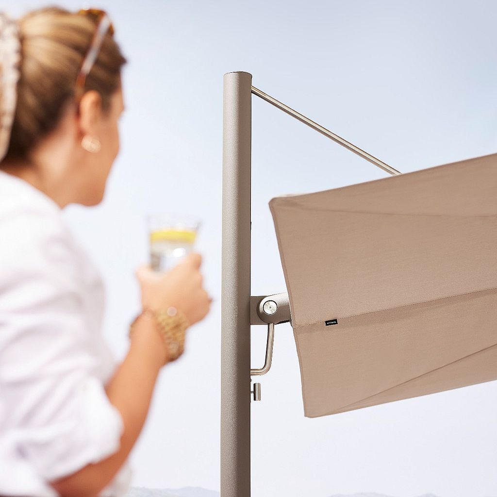 Gros plan sur la toile et le mât métallique d'un parasol Sensu beige. Au premier plan, une femme, un verre à la main, se détache sur un paysage extérieur lumineux.