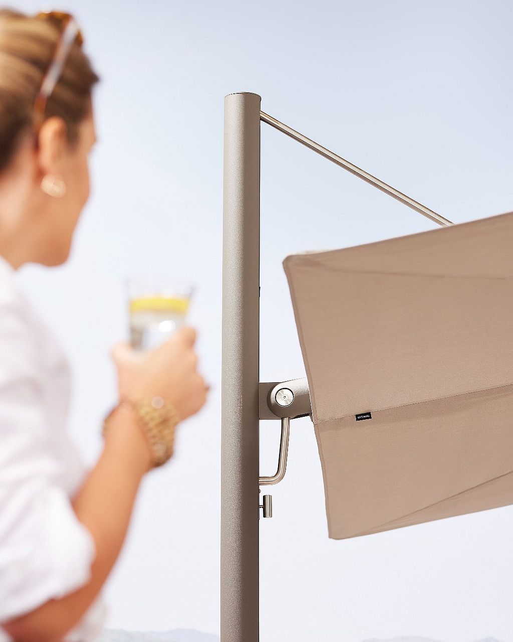 Close-up of a beige Sensu parasol canopy and metal pole, with a woman holding a drink softly blurred in the foreground against a bright outdoor landscape.