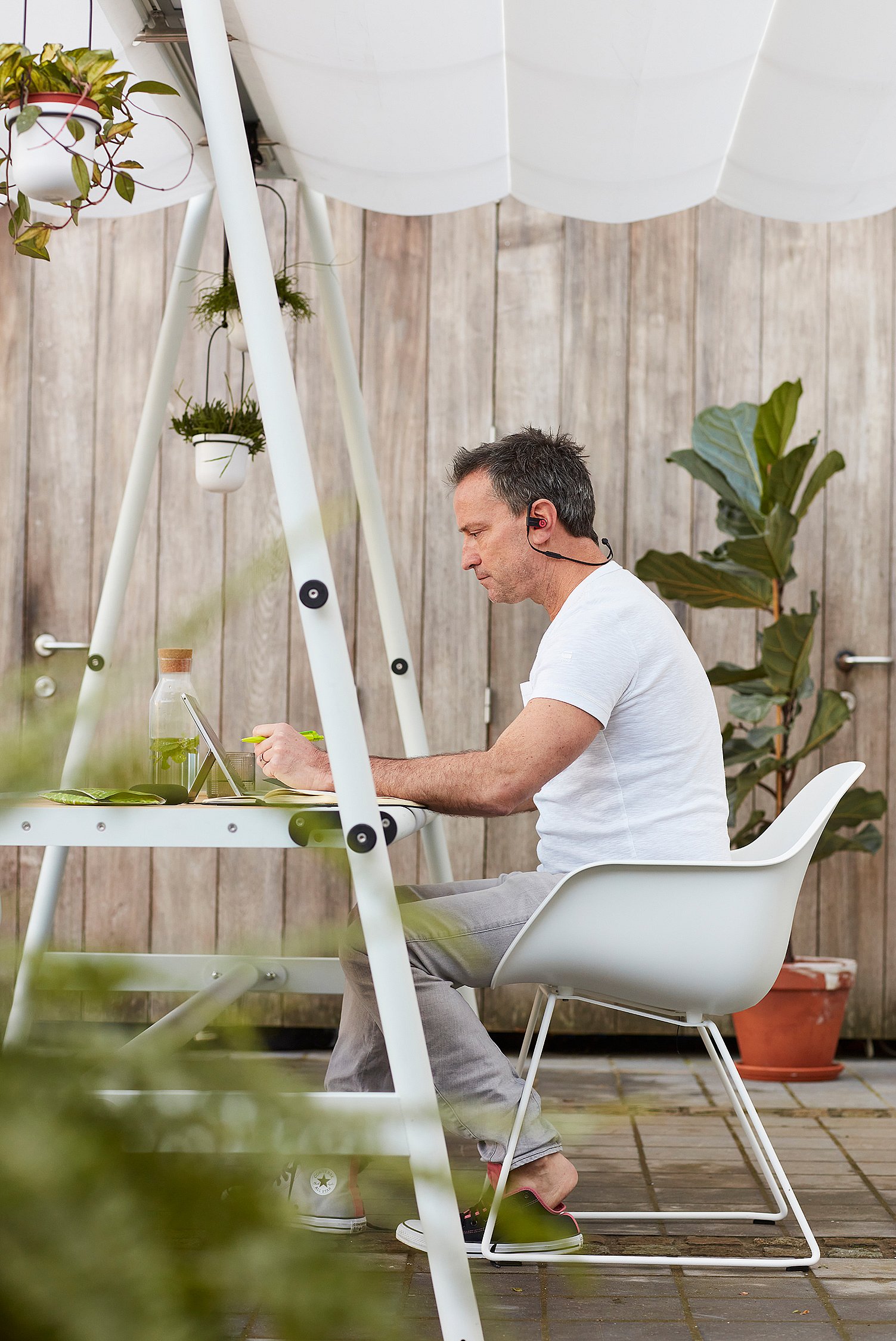 Man wearing a headset works at a tablet under the AMAi shade structure, surrounded by hanging plants and outdoor greenery.