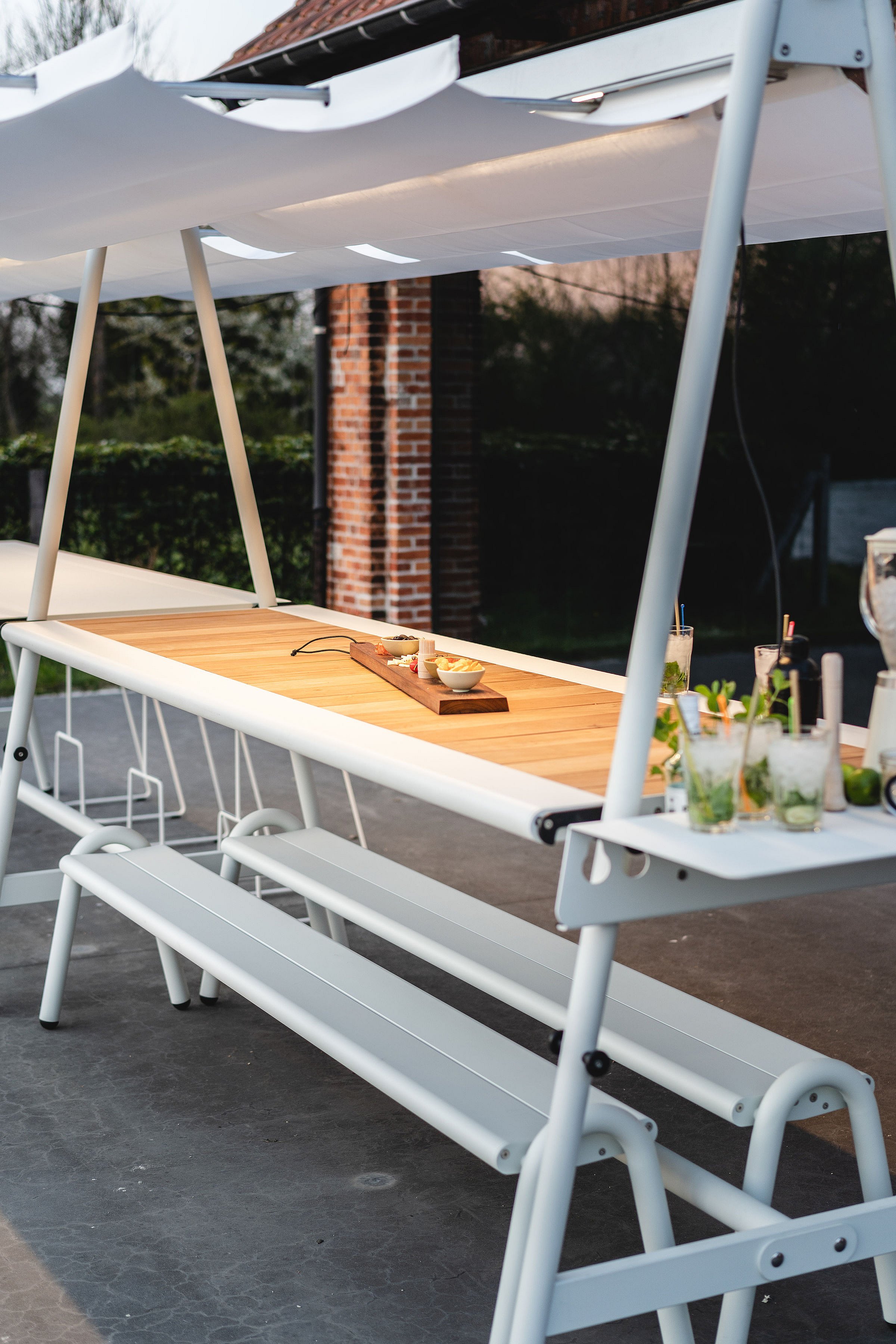 White AMAi table with wooden tabletop, benches, overhead shade, side tray, drinks, and snacks set up on an outdoor terrace.