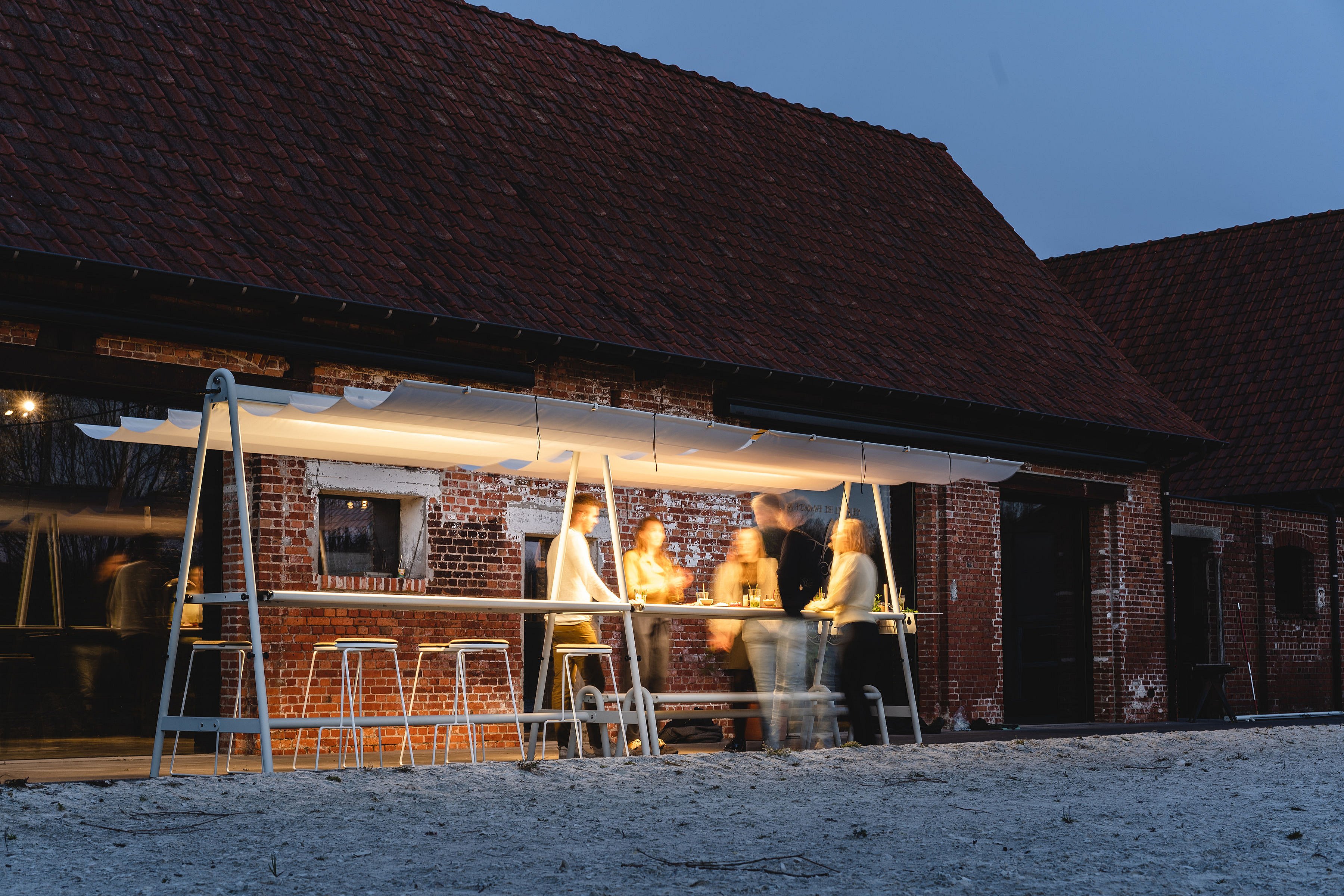 People standing around a lit AMAi table at dusk outside a brick building, with the glowing shade structure creating a warm social atmosphere.