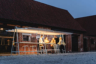 People standing around a lit AMAi table at dusk outside a brick building, with the glowing shade structure creating a warm social atmosphere.