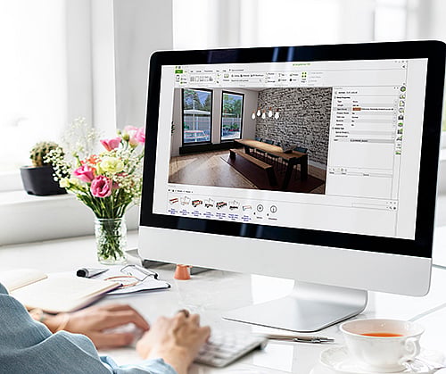 Desktop computer showing the pCon 3D configurator with an Extremis table placed in a virtual interior, while a person works at the desk beside flowers, notes, and a cup of tea.