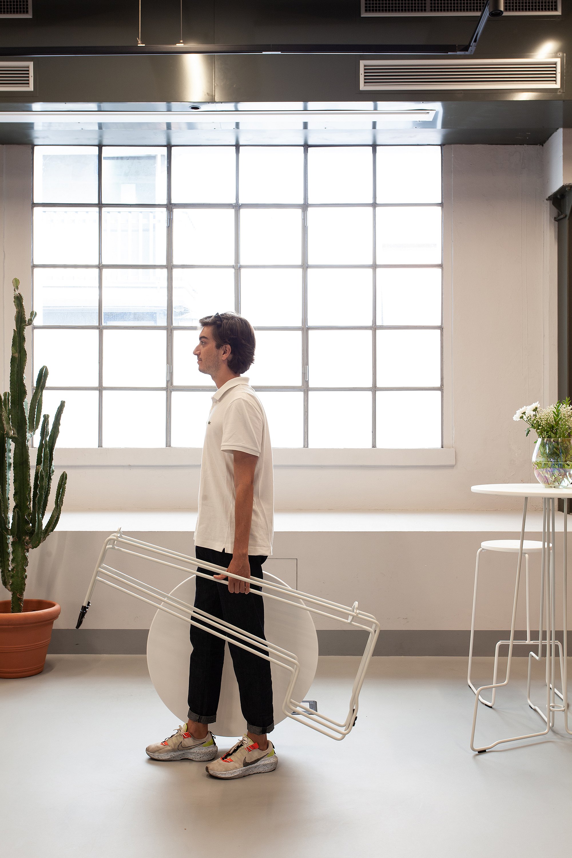   Un homme transporte une table Tiki blanche pliée près d'un grand cactus et d'une fenêtre ; une table Tiki blanche et des tabourets se trouvent à proximité.