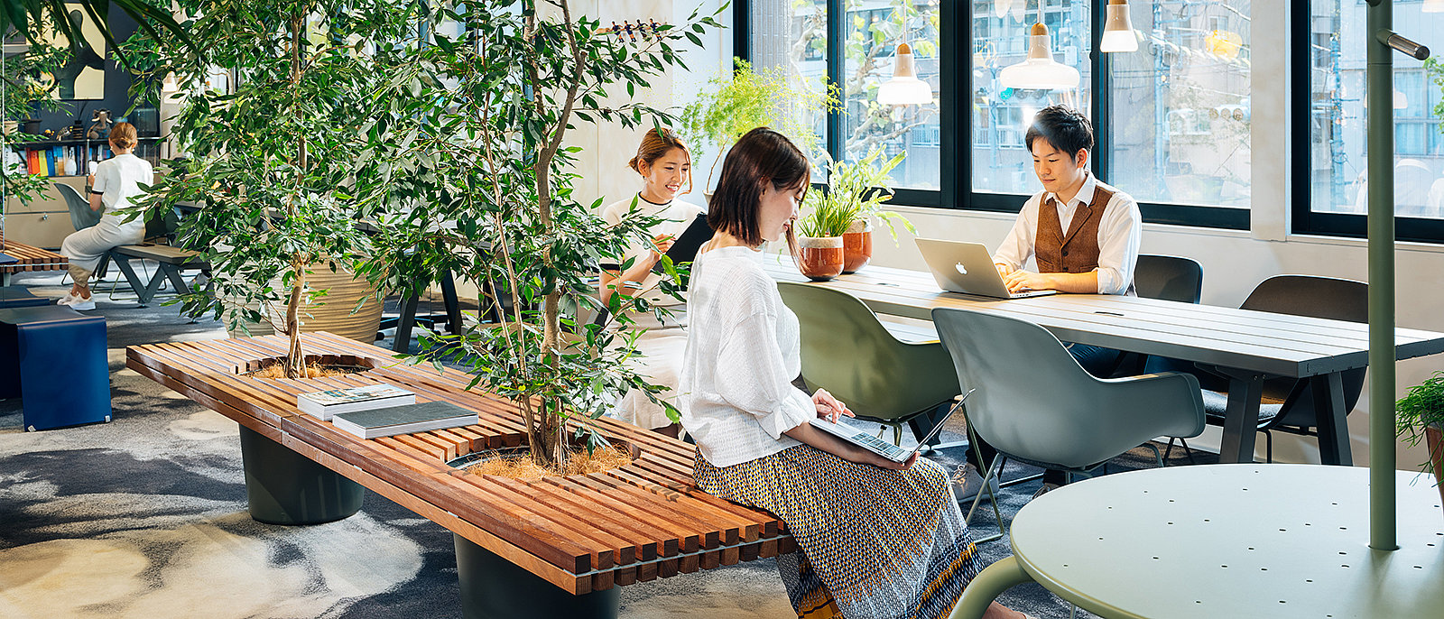   In einem hellen, naturnahen Büro mit Zimmerpflanzen, Sitzbänken, einem langen Gemeinschaftstisch, Hängelampen und großen Fenstern arbeiten und entspannen die Mitarbeiter.