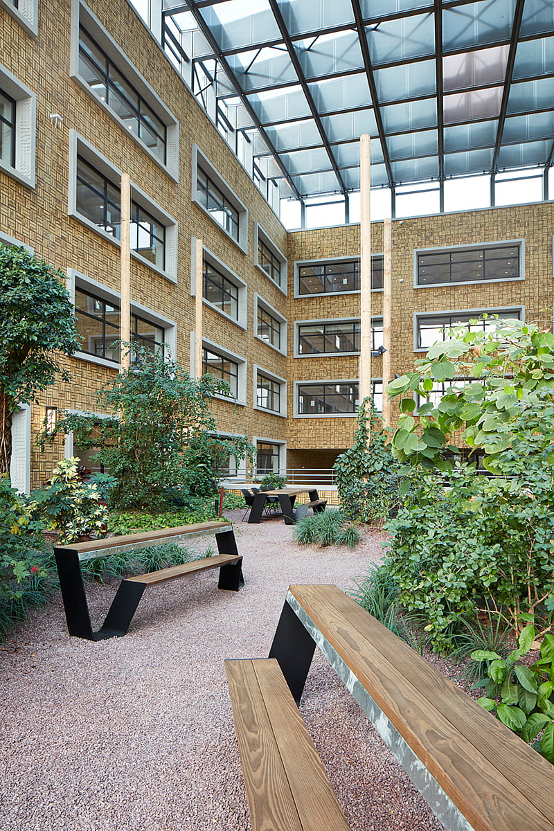Wooden benches and picnic tables arranged in a planted indoor courtyard beneath a glass roof.