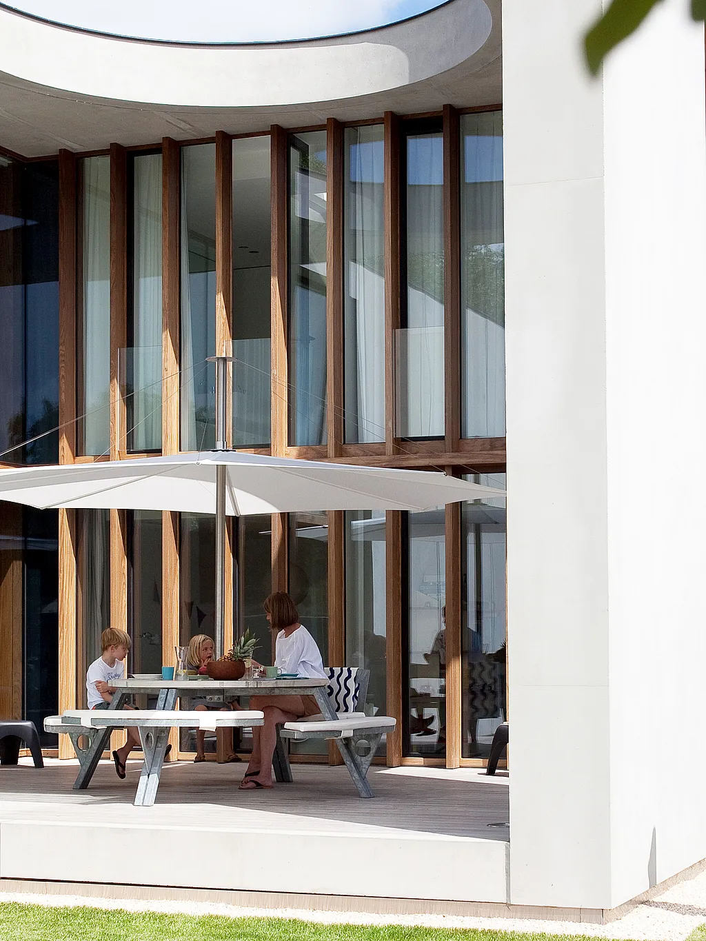 Modern concrete villa with a large covered patio featuring a round Gargantua picnic table and an Inumbra parasol beneath a circular roof opening that lets sunlight illuminate the terrace.