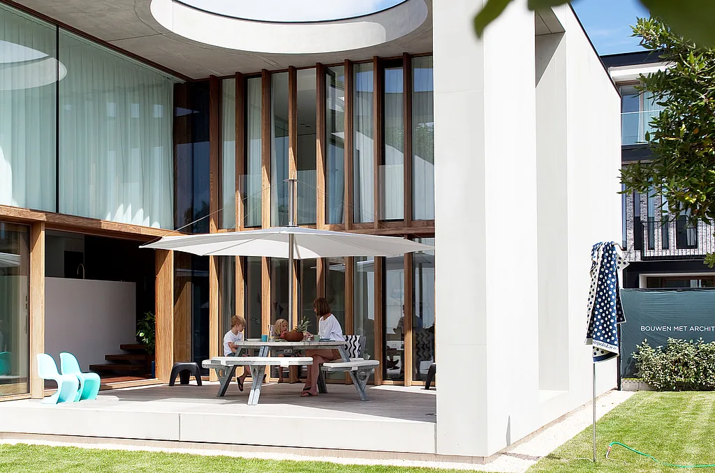 Villa moderne en béton avec une grande terrasse couverte équipée d'une table de pique-nique ronde Gargantua et d'un parasol Inumbra, sous une ouverture circulaire dans le toit laissant entrer la lumière du soleil.