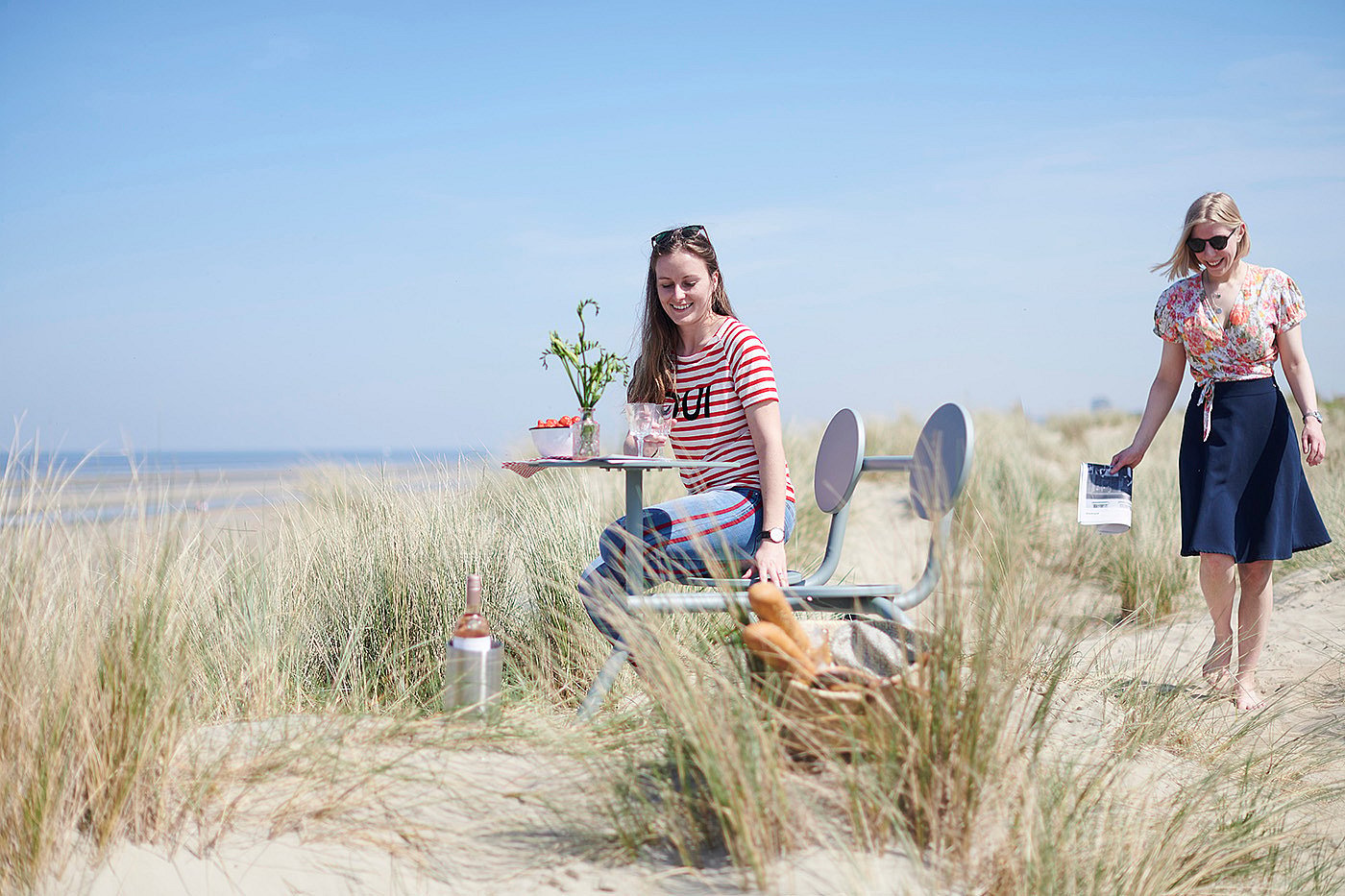 Two women setting up a light blue Bistroo table in the sand dunes with picnic items and sea views