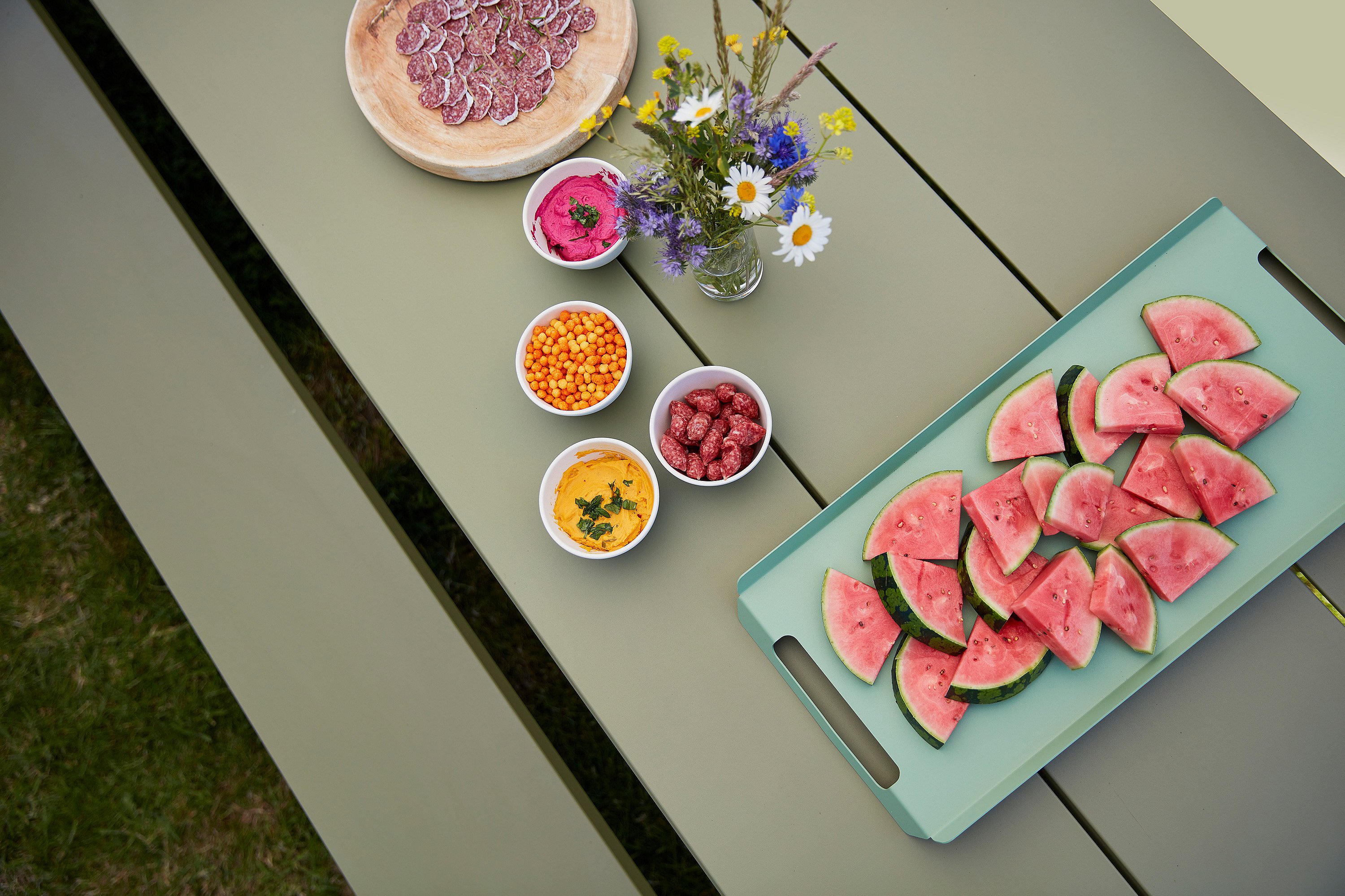 Wassermelonenscheiben, Dips, Beeren und Blumen auf einem grünen Hopper-Picknicktisch angeordnet