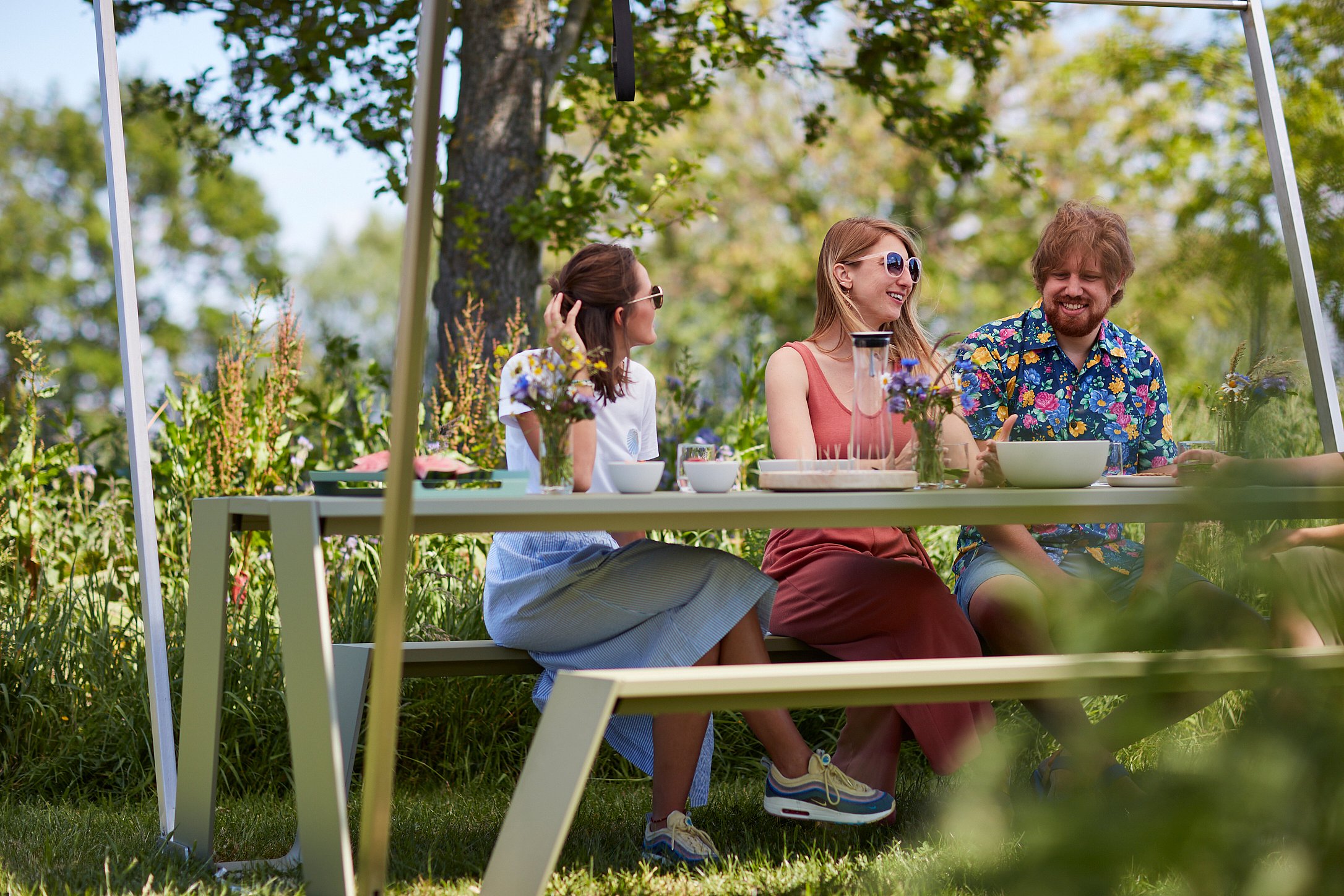 Menschen lächeln um einen Hopper-Picknicktisch mit Picknickessen und Blumen im Schatten