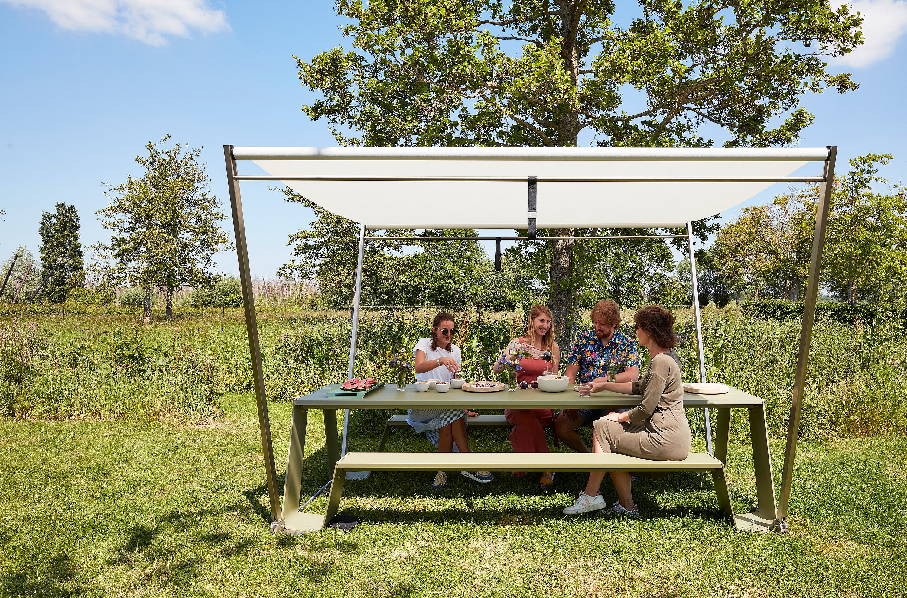 Een gezin van vier eet aan een groene Hopper picknicktafel onder een witte Hopper parasol in een grasveld.