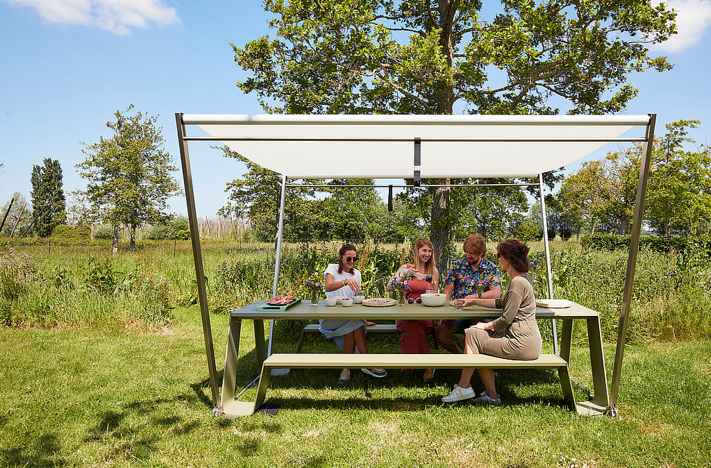 Een gezin van vier eet aan een groene Hopper picknicktafel onder een witte Hopper parasol in een grasveld.