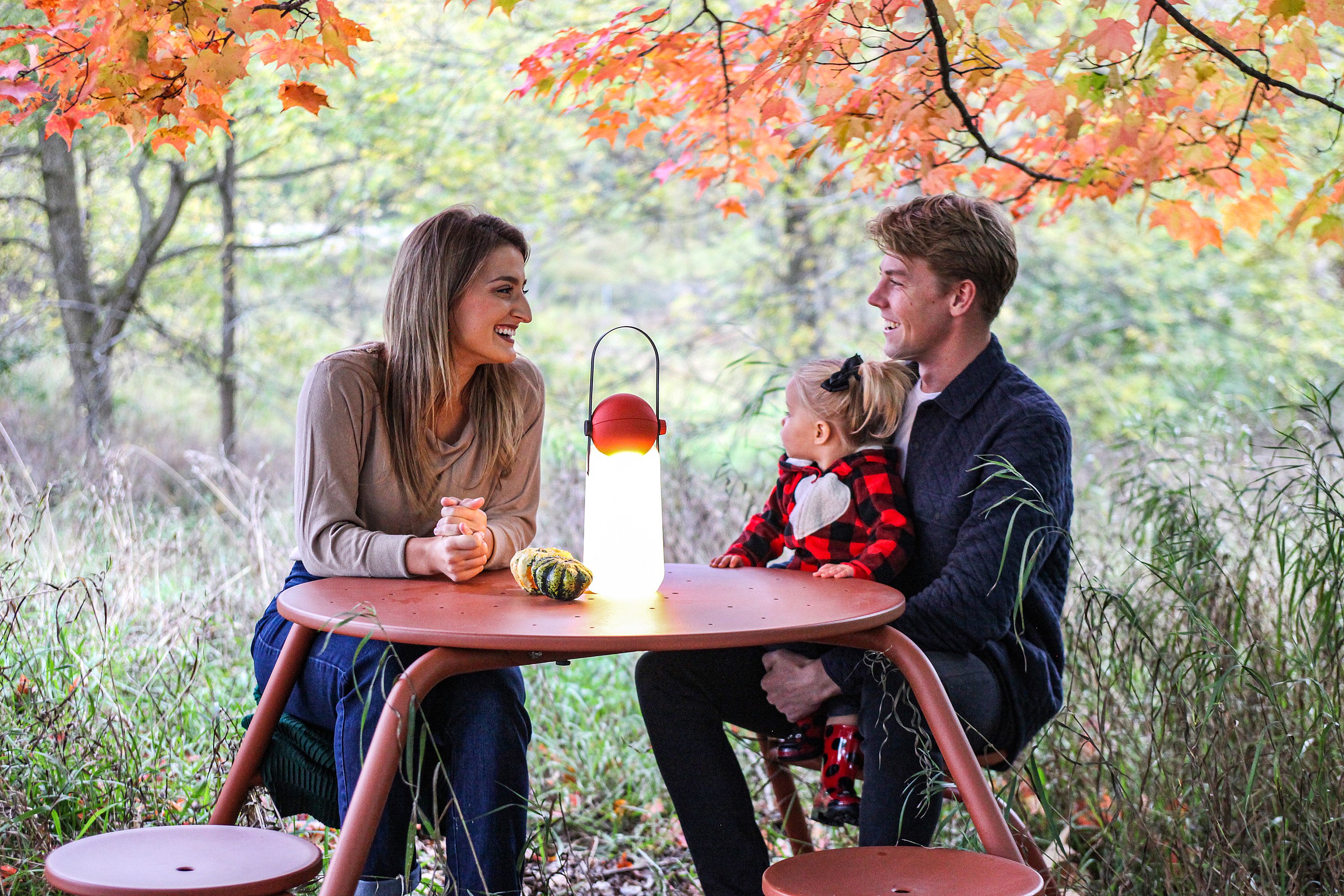 Familie sitzt um einen kupferbraunen Virustisch mit beleuchteter Tischlampe unter orangefarbenen Herbstblättern