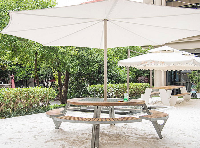 Table ronde Gargantua avec des bancs en bois sous une teinte Inumbra sur la terrasse d’un café inspiré de la plage